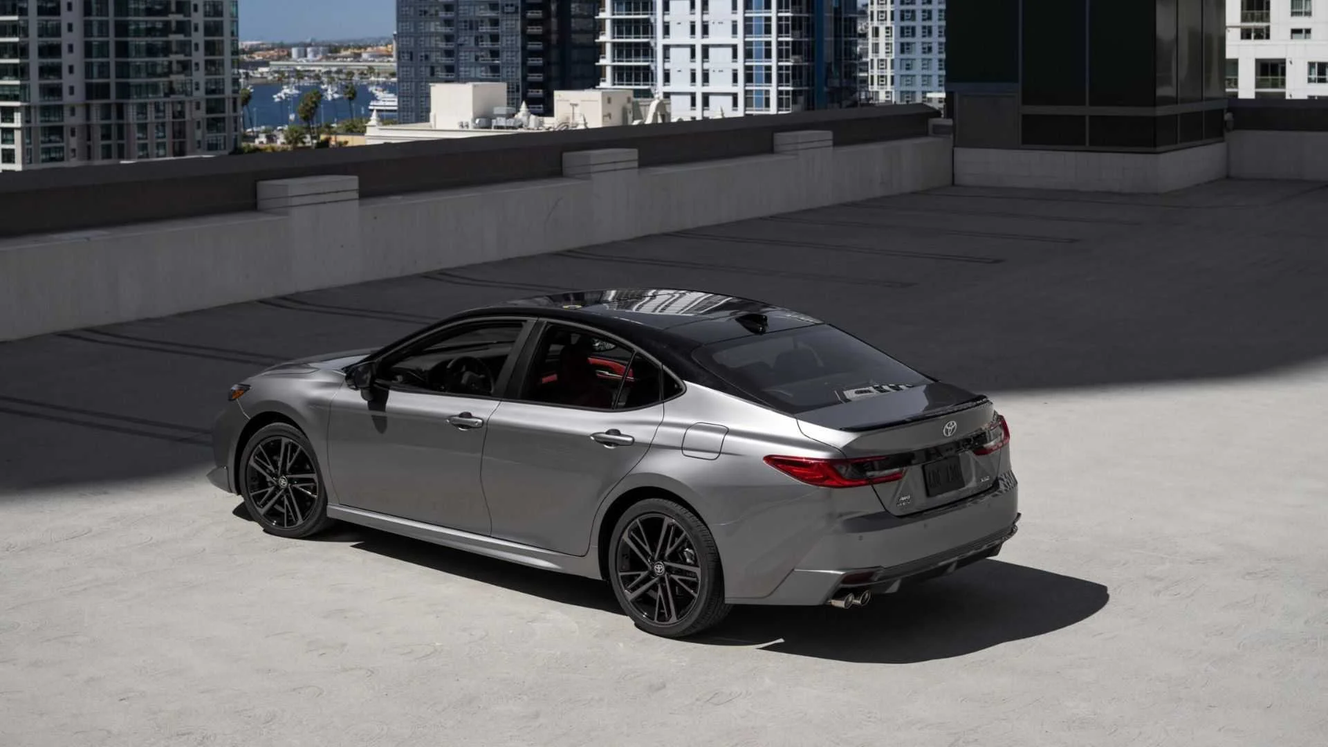 Static rear 3/4 shot of a gray 2025 Toyota Camry XSE AWD parked with highrise buildings in the background.