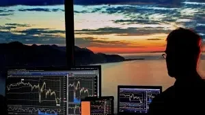Silhouette of a person looking at multiple financial trading screens with charts against a sunset landscape.